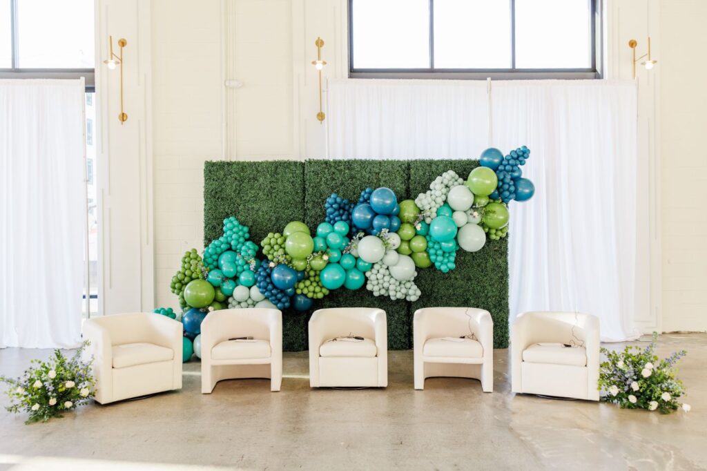 A vibrant green balloon wall with a sprawling organic arch of blue, green, and teal balloons, set behind four white upholstered chairs arranged for an event or panel discussion. Two floral arrangements with white and purple flowers flank the chairs. The setting features white curtains and tall windows in a bright, modern space.