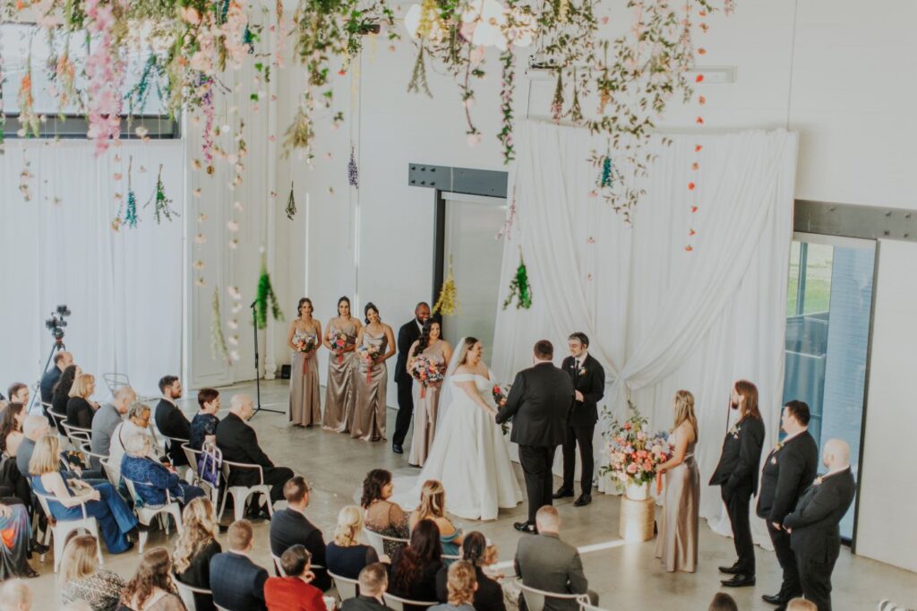 Guests gather for an indoor wedding ceremony at a Charlotte, NC event venue, featuring a modern aesthetic with flowing white drapes and abundant hanging floral arrangements. The bride and groom stand at the altar with their wedding party, while attendees are seated in rows.