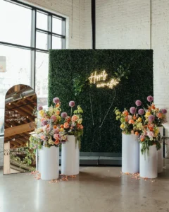 Bright spring florals on white pedestals stood in front of a large hedge wall with neon sign reading happily ever after and a 'welcome to our wedding' sign on a large stand up mirror