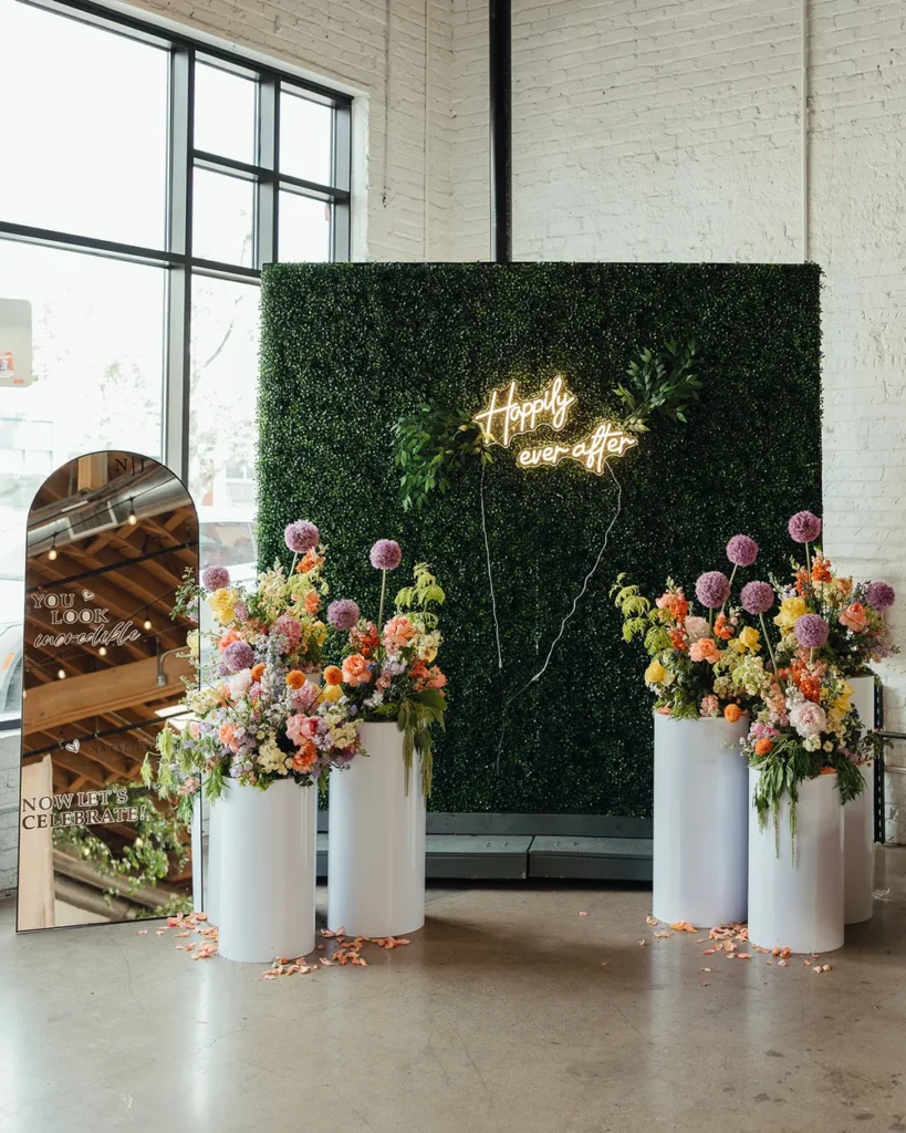 Bright spring florals on white pedestals stood in front of a large hedge wall with neon sign reading happily ever after and a 'welcome to our wedding' sign on a large stand up mirror