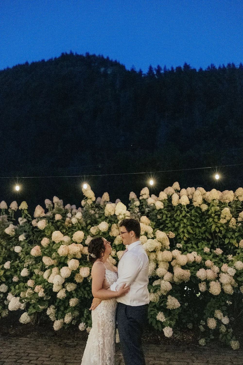 Bride and groom standing in front of a large bush of flowers at night